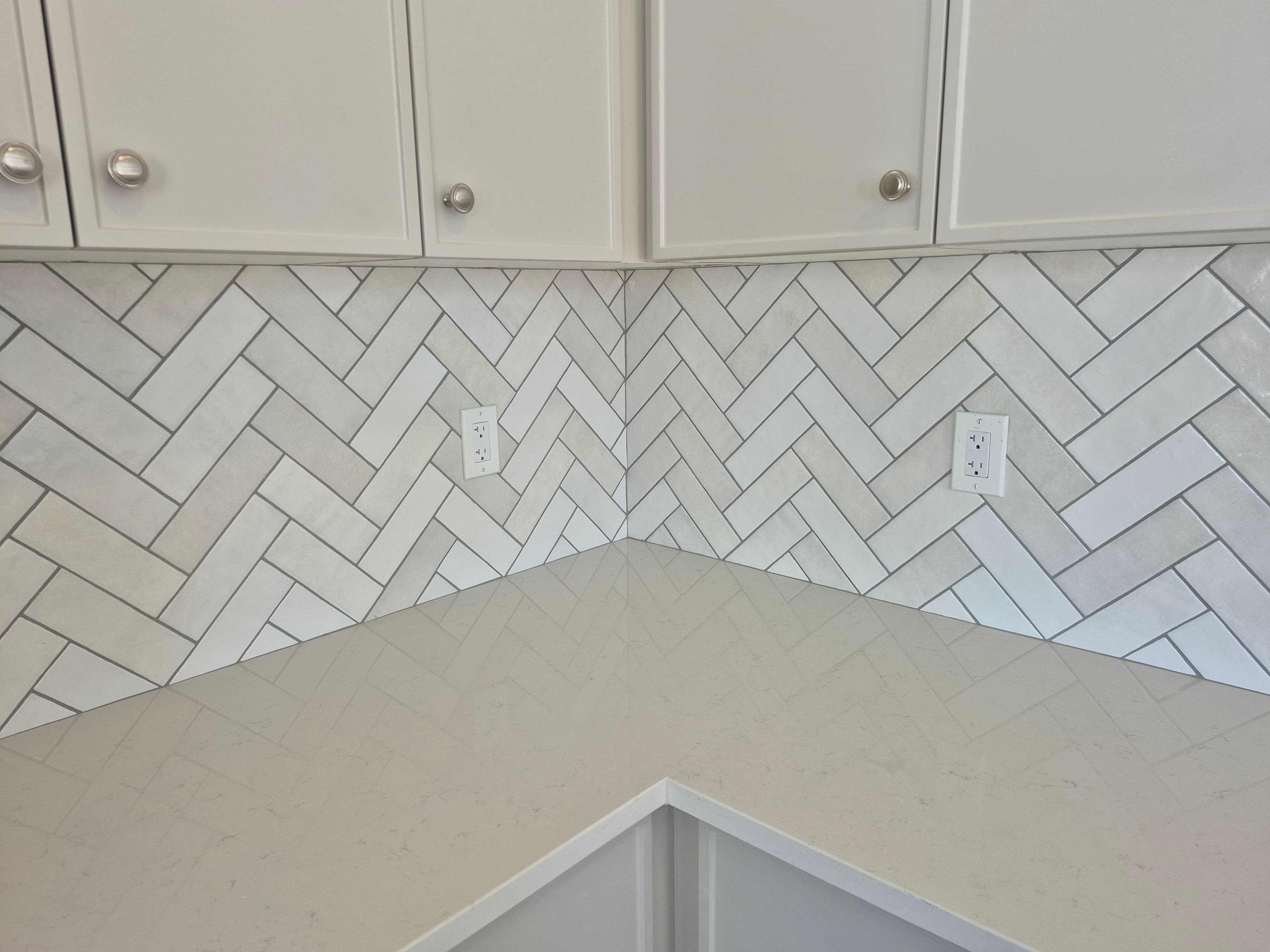 White kitchen corner featuring a herringbone tile backsplash, light quartz countertops, and white cabinetry.