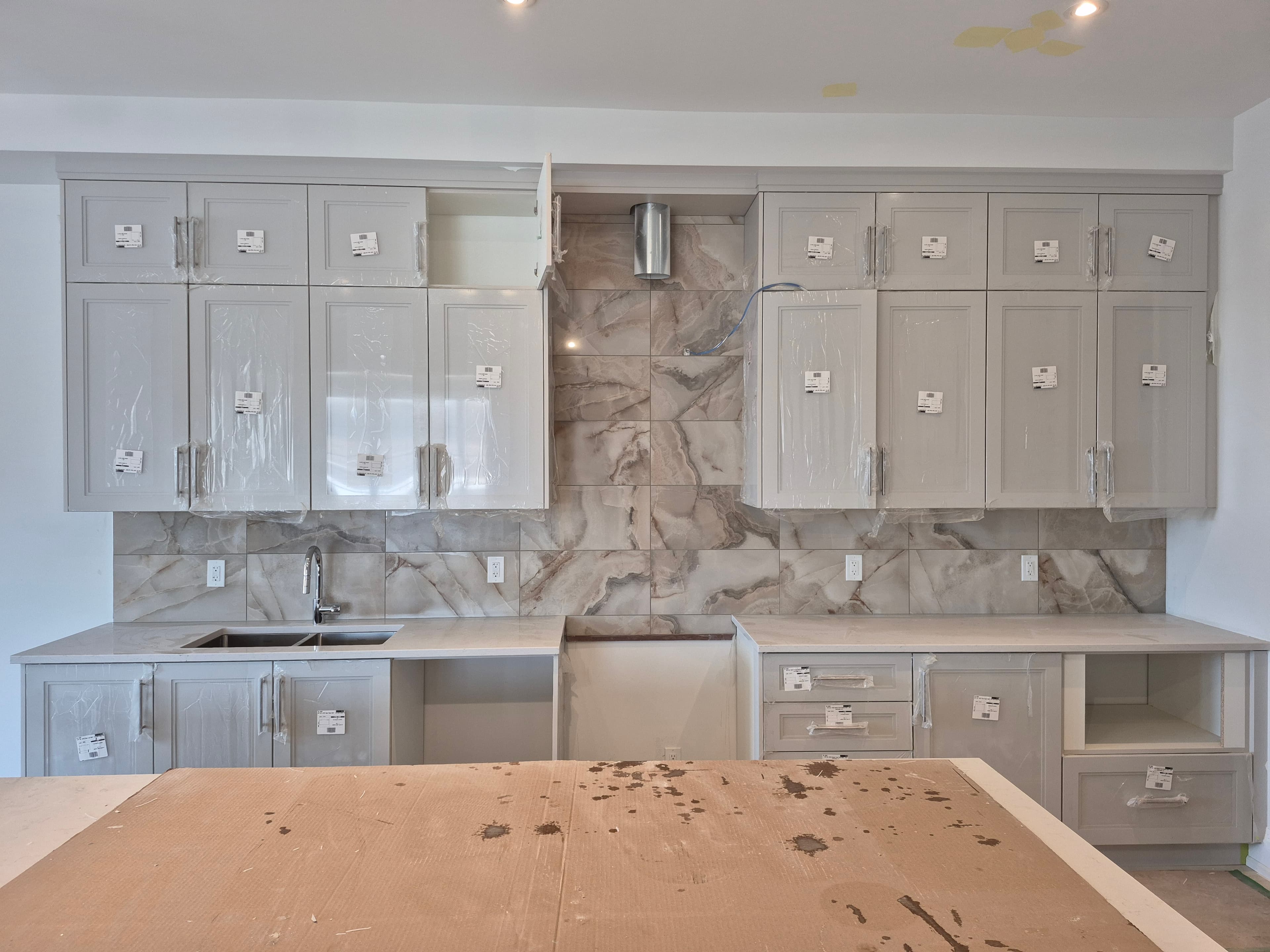 Modern kitchen under construction featuring grey cabinets, marble backsplash, and a cardboard-covered island.